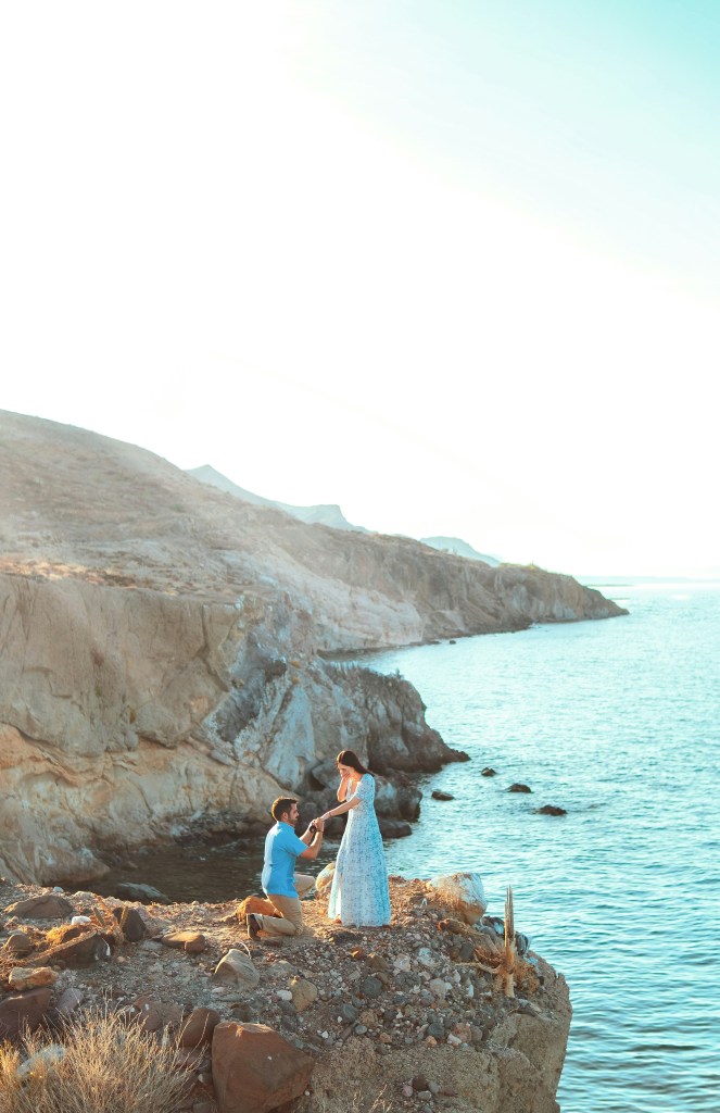 novio arrodillado pidiendo matrimonio junto a mujer en una formacion rocosa cerca del mar